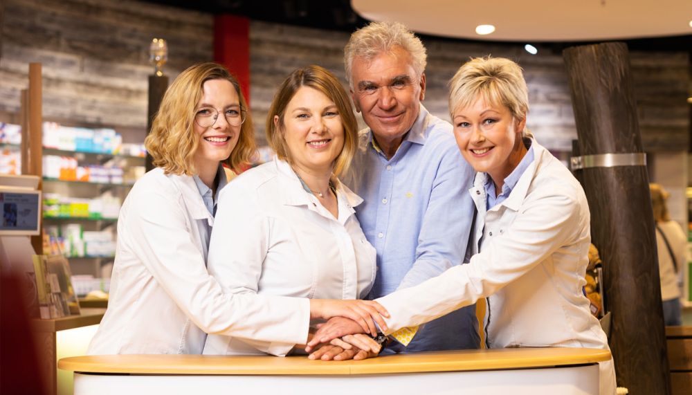 Apothekenteam steht geschlossen hinter dem HV-Tisch und legt die Hände zusammen